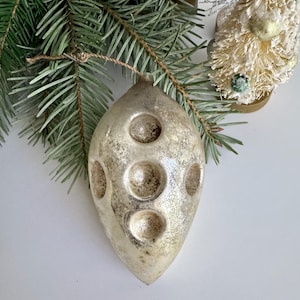 May include: A vintage silver Christmas ornament with a textured surface and circular indentations. The teardrop-shaped ornament hangs from a pine branch, suggesting a festive holiday theme. A small, white bottle brush tree is in the background.