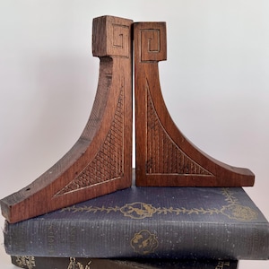 May include: Pair of dark brown wooden bookends with a geometric design. Each bookend has a square top with a carved spiral and a curved body with a textured pattern. The bookends are resting on top of a stack of vintage books.