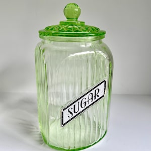 May include: A light green, translucent glass canister with vertical ribbing and a matching lid featuring a knob handle. A white, rectangular label with a black border and the word SUGAR in black, stylized capital letters is attached to the front.