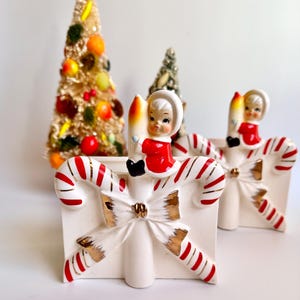May include: Vintage Christmas candle holders with candy cane and bow details. Each holder features a small figurine in a red outfit holding a candle. Two decorative Christmas trees are in the background. The candle holders are white with red and gold accents.