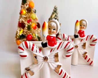 May include: Vintage Christmas candle holders with candy cane and bow details. Each holder features a small figurine in a red outfit holding a candle. Two decorative Christmas trees are in the background. The candle holders are white with red and gold accents.