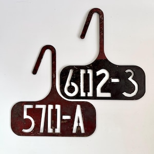 May include: Two vintage metal tags with a hanger-like design. One tag reads "570-A" in white, the other reads "6112-3". Both tags have a dark red-brown color with a distressed finish, suggesting age and use.