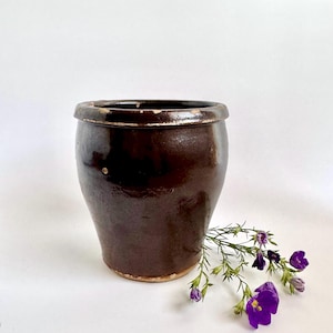 May include: A dark brown ceramic pot with a slightly flared shape and a rounded rim. The pot has a glossy finish and a few small imperfections. A sprig of purple flowers rests near the pot, adding a touch of color.
