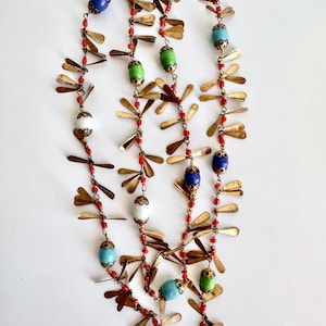 May include: A multi-strand necklace featuring colorful beads in blue, green, white, and turquoise, interspersed with small red beads and gold-colored metal accents. The necklace has a unique, layered design.