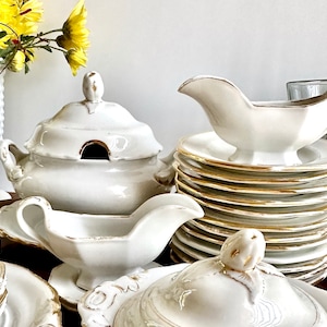May include: A set of white porcelain dinnerware with gold trim. The set includes a large serving bowl with a lid, two sauce boats, and a stack of plates.