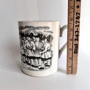 May include: Cream-colored ceramic mug with a black and white illustration of a group of people wearing hats and coats. The mug has a curved handle and is next to a wooden ruler.
