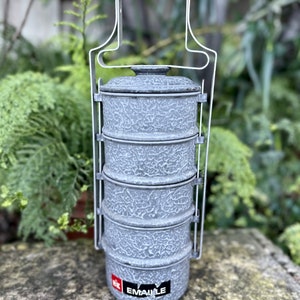 May include: A vintage gray enamel lunch carrier with four stacked compartments and a metal handle. The carrier has a textured surface and a black handle. The word "EMAILLE" is visible on a sticker on the bottom.