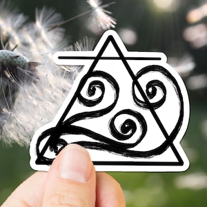 May include: A black and white sticker with a triangle shape and a swirling design inside. The sticker is being held by a hand against a blurry background of dandelion seeds.