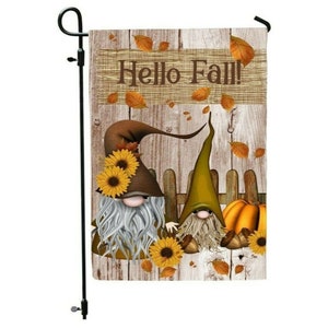 May include: A garden flag with a rustic wood background featuring two gnomes, one with a brown hat and the other with a green hat. The gnomes are sitting in front of a wooden fence with sunflowers and fall leaves around them. The text "Hello Fall!" is written on a wooden sign above the gnomes.