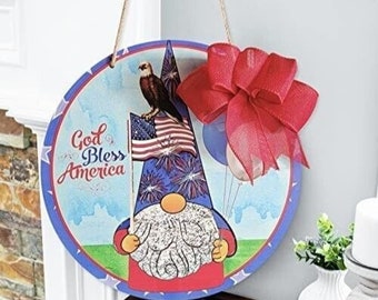 May include: A round wooden door hanger with a gnome wearing a red, white, and blue hat and holding an American flag. The gnome is surrounded by fireworks and the words "God Bless America" are printed on the hanger. A red bow is attached to the top of the hanger.