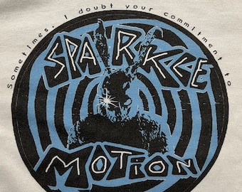 May include: White t-shirt with a blue and black circular graphic. The graphic features a black and white illustration of a creature with a star on its forehead. The text "Sometimes I doubt your commitment to SPARKLE MOTION" is written around the graphic.