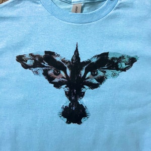 Könnte beinhalten: Ein hellblaues T-Shirt mit einem schwarzen und weißen Grafikdruck eines Vogels mit großen Augen.