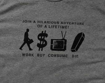 May include: Gray t-shirt with a black graphic design. The design includes a silhouette of a person, a dollar sign, a television, and a coffin. Text reads "JOIN A HILARIOUS ADVENTURE OF A LIFETIME! WORK BUY CONSUME DIE."
