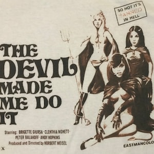 Puede incluir: Una camiseta blanca con el póster de la película vintage "The Devil Made Me Do It" con tres mujeres en trajes de diablo. El texto "So Hot It's Banned In Hell" y "Eastmancolor" está impreso en la parte inferior de la camiseta.