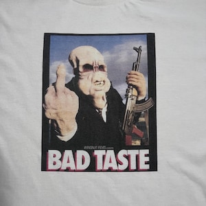 May include: A white t-shirt featuring a graphic print of a pale, alien-like creature in a black suit, holding a rifle and giving the middle finger. Below the image, the text reads WINGNUT FILMS presents BAD TASTE in bold white and pink letters.