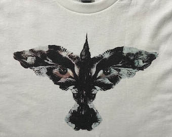 May include: White t-shirt with a black and white graphic print of a bird with large eyes. The bird is stylized to look like a raven or crow.