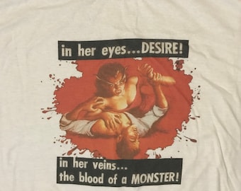 May include: White t-shirt featuring a graphic print of a woman with fangs biting a man's neck. The graphic is surrounded by red splatter paint. The text on the shirt reads "in her eyes...DESIRE!" and "in her veins...the blood of a MONSTER!"
