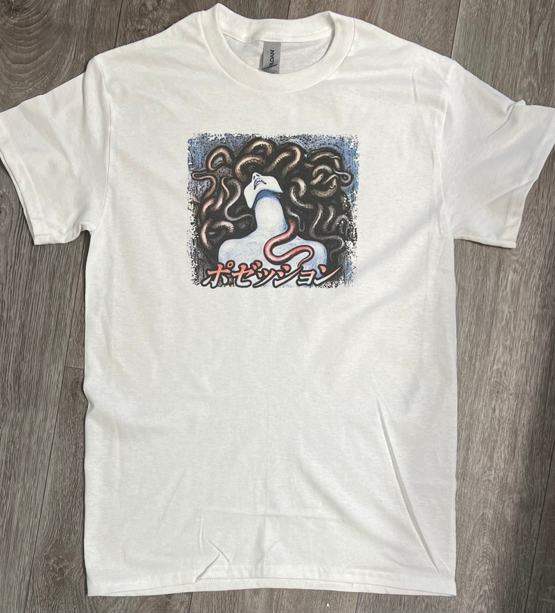 Puede incluir: Camiseta blanca con un estampado gr&aacute;fico de una mujer con serpientes como cabello. El texto "ホセダンジョン" est&aacute; impreso debajo de la imagen.