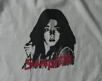 May include: A white crew neck t-shirt featuring a black and white graphic print of a person with long dark hair holding a sharp object. The word Suspiria is printed in bold red letters across the bottom of the graphic design on the front.