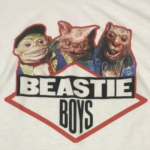 May include: White t-shirt with a red diamond-shaped graphic featuring the text "BEASTIE BOYS" in black and three cartoon characters with green, pink, and brown skin.