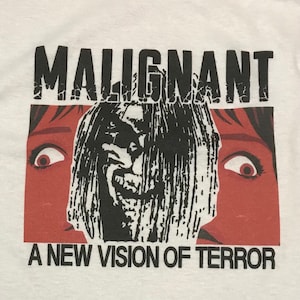Puede incluir: Camiseta blanca con un estampado gráfico en blanco y negro de una cara aterradora con el texto "MALIGNANT" y "A NEW VISION OF TERROR" impreso debajo.