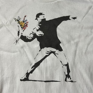 Könnte beinhalten: Ein weißes T-Shirt mit einem Schwarzweißbild einer Person, die einen Blumenstrauß wirft. Die Person trägt eine Maske und hat den Arm ausgestreckt.