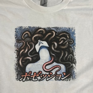 Puede incluir: Camiseta blanca con un estampado gr&aacute;fico de una mujer con serpientes como cabello. El texto "オセッショ" est&aacute; impreso debajo de la imagen.