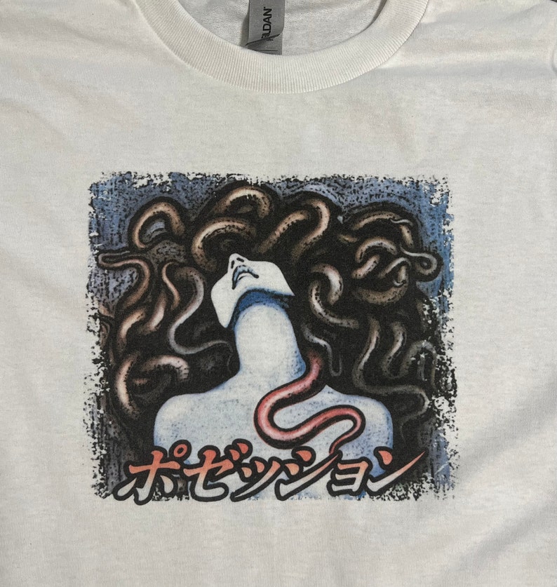 Puede incluir: Camiseta blanca con un estampado gr&aacute;fico de una mujer con serpientes como cabello. El texto "オセッショ" est&aacute; impreso debajo de la imagen.
