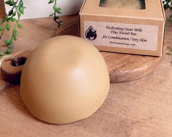 May include: A smooth, round, tan-coloured facial bar soap next to a square cardboard box with a clear window. The box label reads: Hydrating Goat Milk Clay Facial Bar for Combination/Dry Skin and includes the website TheYummySoap.com.