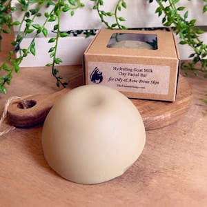 May include: A brown cardboard box with a clear window showing a green clay facial bar. The box has a white label with the text "Hydrating Goat Milk Clay Facial Bar for Oily & Acne Prone Skin TheYummySoap.com". A green clay facial bar is sitting on a wooden surface.