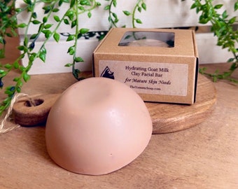 May include: A dome-shaped, light pink facial bar sits in front of a brown cardboard box with a clear window. The label on the box reads Hydrating Goat Milk Clay Facial Bar for Mature Skin Needs and includes the website TheYummySoap.com.