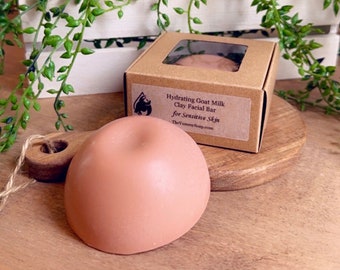 May include: A brown cardboard box with a clear window showing a light brown clay facial bar. The box has a white label with the text "Hydrating Goat Milk Clay Facial Bar for Sensitive Skin TheYummySoap.com". The bar is sitting on a wooden chopping board.