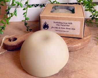 May include: A smooth, dome-shaped, light beige facial soap bar sits in front of a square brown cardboard box with a clear window. The label reads: Hydrating Goat Milk Clay Facial Bar for Oily & Acne Prone Skin and TheYummySoap.com.