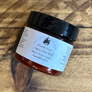 Puede incluir: Un pequeño frasco marrón con tapa negra que contiene Breathe Easy Vapor Chest Rub, un producto natural y holístico. La etiqueta presenta un logotipo azul y blanco con un pájaro estilizado y el texto "Breathe Easy Vapor Chest Rub" en blanco. El frasco está sobre una superficie de madera.