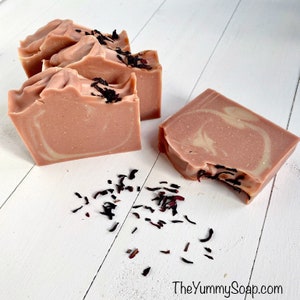 Puede incluir: Cuatro barras de jabón hecho a mano con un patrón de remolino rosa y blanco. El jabón está decorado con flores de hibisco secas. El jabón está sobre una superficie de madera blanca.