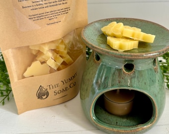 Może przedstawiać: Brązowa papierowa torebka z etykietą Natural Beeswax Melts i przezroczystym okienkiem ukazującym żółte kształty wosku, obok zielonego ceramicznego kominka zapachowego ze świeczką typu tealight i żółtymi woskowymi motylkami na górze.