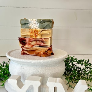 May include: A stack of four rectangular soap bars with varied textures and colors, including sage green with white flecks, beige with gold crystals, pink and white swirls with dried petals, and orange with a visible cinnamon stick embedded.