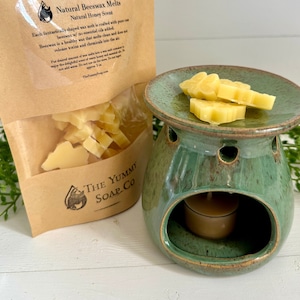 May include: A brown paper bag with a clear window showing yellow beeswax melts. The bag has a label that reads "Natural Beeswax Melts Natural Honey Scent". The bag is next to a green ceramic wax warmer with a small tea light candle inside. The wax warmer has a small dish on top with more yellow beeswax melts.