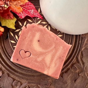 May include: A rectangular bar of soap in a dusty rose color with a marbled texture and a small heart shape stamped into the lower left corner. The soap has a smooth surface and is positioned on a dark brown circular wooden base.