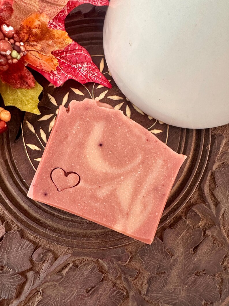 May include: A rectangular bar of soap in a dusty rose color with a marbled texture and a small heart shape stamped into the lower left corner. The soap has a smooth surface and is positioned on a dark brown circular wooden base.