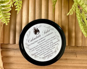 May include: A round black tin of Calendula Salve with a white label. The label features a logo, the text 'Calendula Salve', usage instructions for bug bites and skin irritations, a list of ingredients including olive oil and beeswax, and '2 oz'.
