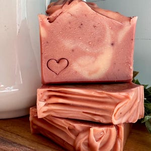 May include: A stack of three rectangular bars of soap in a dusty rose color with cream-colored swirls. The top bar features a small, stamped heart outline on the front face. The soap has a smooth, matte texture with visible fine particles.