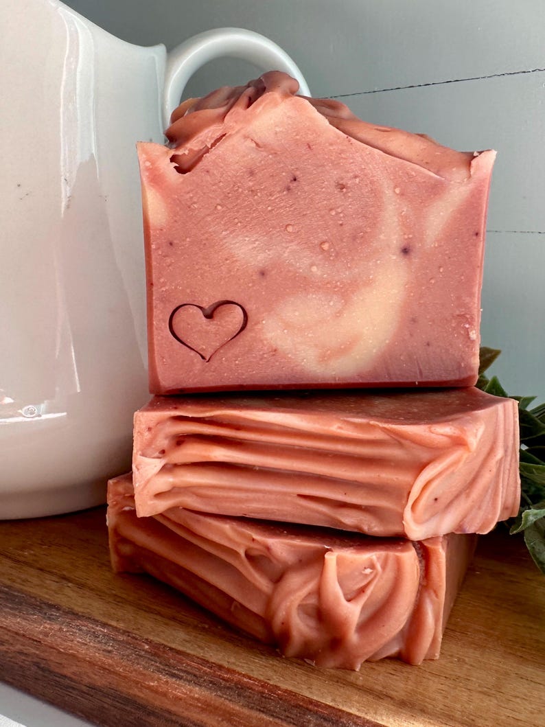 May include: A stack of three rectangular bars of soap in a dusty rose color with cream-colored swirls. The top bar features a small, stamped heart outline on the front face. The soap has a smooth, matte texture with visible fine particles.