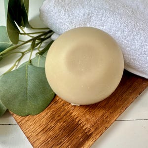 May include: A round, light beige soap bar rests on a small, angled wooden tray, next to a folded white towel. Eucalyptus leaves and stems are arranged on the left side. The soap has a smooth surface and a slight indentation on top, suggesting it's handmade.
