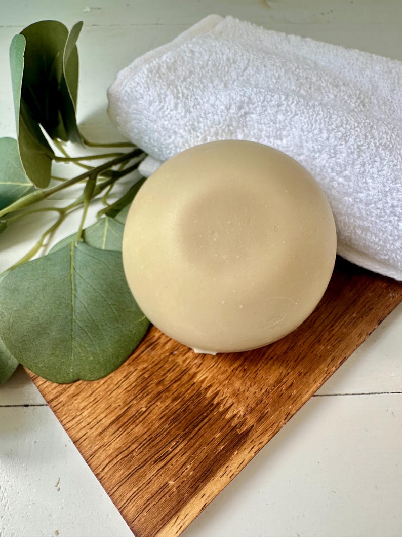 May include: A round, light beige soap bar rests on a small, angled wooden tray, next to a folded white towel. Eucalyptus leaves and stems are arranged on the left side. The soap has a smooth surface and a slight indentation on top, suggesting it's handmade.