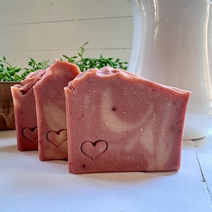 May include: Three rectangular bars of pink soap with a marbled white swirl pattern. Each bar features a small, stamped heart outline on the front surface. The soap has a textured, uneven top edge and a smooth, solid body with visible speckles.