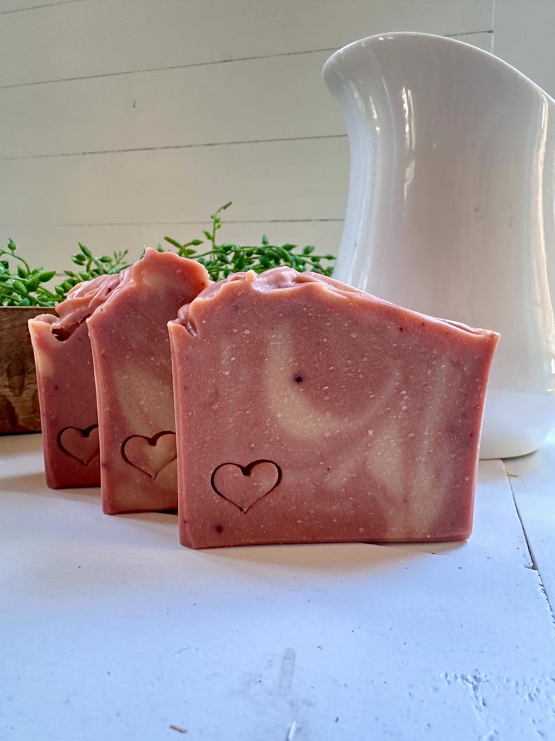May include: Three rectangular bars of pink soap with a marbled white swirl pattern. Each bar features a small, stamped heart outline on the front surface. The soap has a textured, uneven top edge and a smooth, solid body with visible speckles.