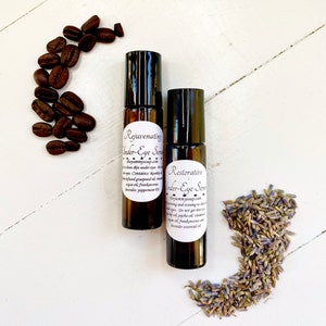 May include: Two amber glass roller bottles with black caps and white labels. The labels read "Rejuvenating Under-Eye Serum" and "Restorative Under-Eye Serum". The bottles are surrounded by coffee beans and lavender flowers.