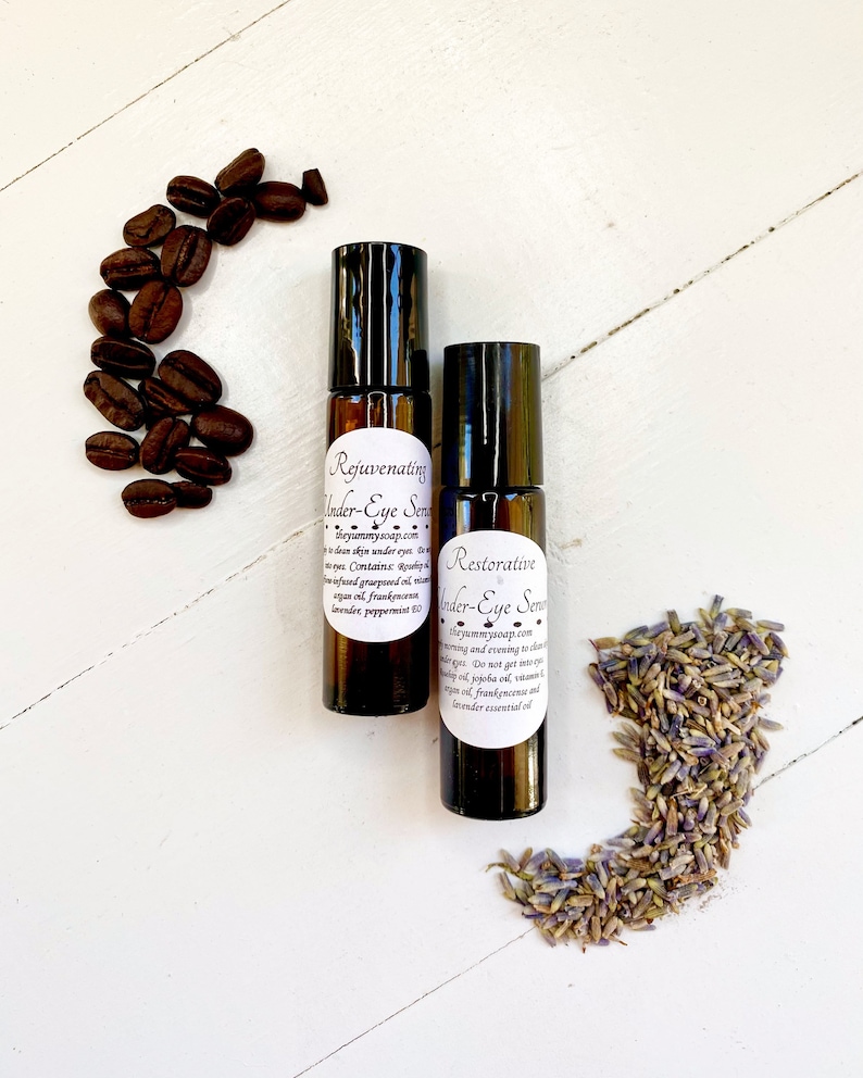 May include: Two amber glass roller bottles with black caps and white labels. The labels read "Rejuvenating Under-Eye Serum" and "Restorative Under-Eye Serum". The bottles are surrounded by coffee beans and lavender flowers.