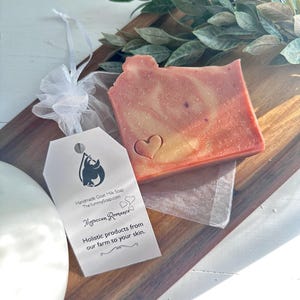 May include: A rectangular bar of handmade goat milk soap with a marbled pink and orange hue, featuring an embossed heart. A white tag attached with a ribbon displays the text: Handmade Goat Milk Soap, TheYummySoap.com, Moroccan Romance.